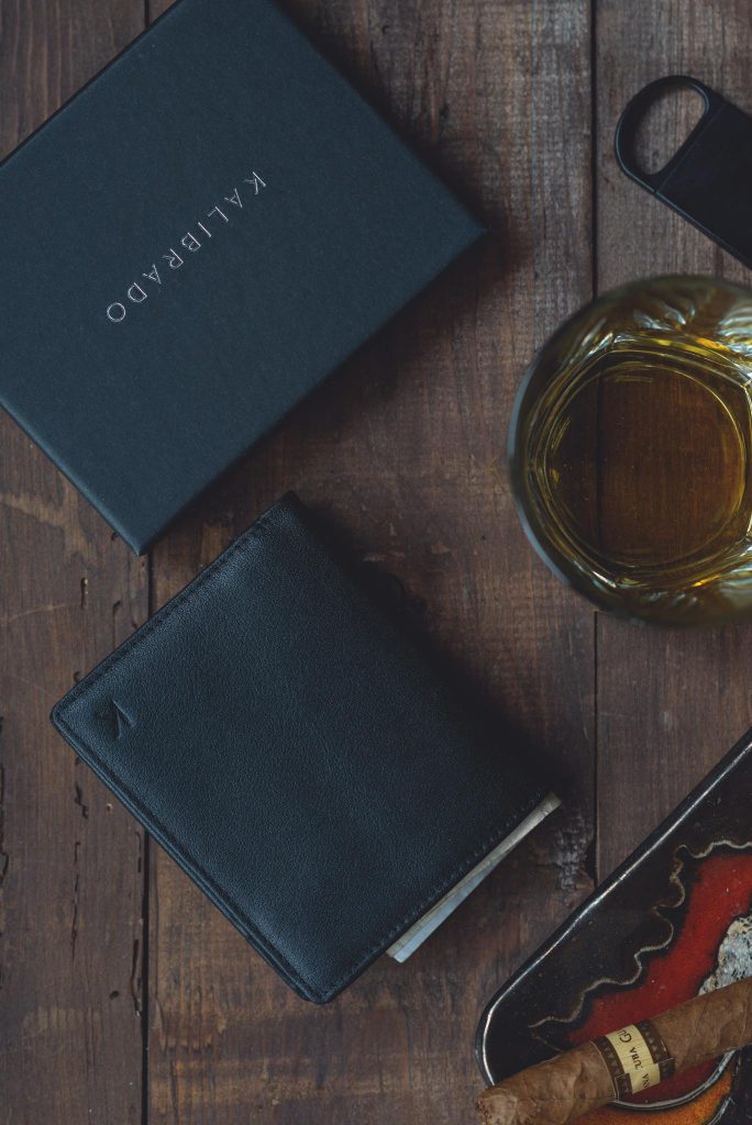Sophisticated arrangement featuring a leather wallet, whiskey glass, and cigar on wooden surface.