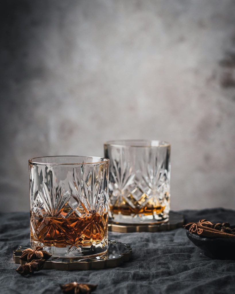Two crystal whiskey glasses with amber liquid on a textured surface, evoking sophistication.